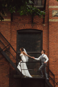 Mariés se tenant les mains en montant des escaliers, Séance photos de mariage dans un espace urbain avec un mur en brique rouge, Mariage à Montréal à l'Espace Canal