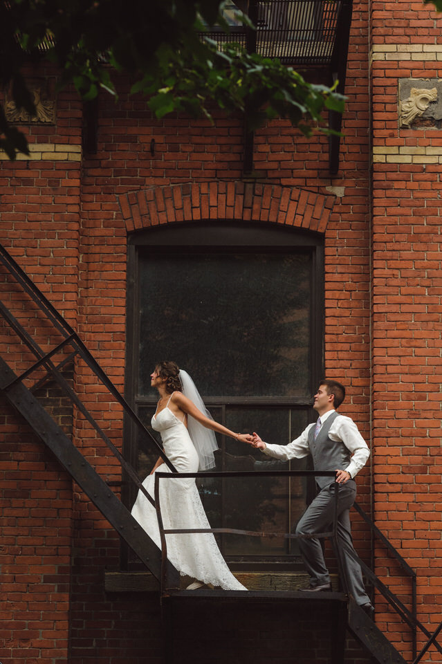 Mariés se tenant les mains en montant des escaliers, Séance photos de mariage dans un espace urbain avec un mur en brique rouge, Mariage à Montréal à l'Espace Canal